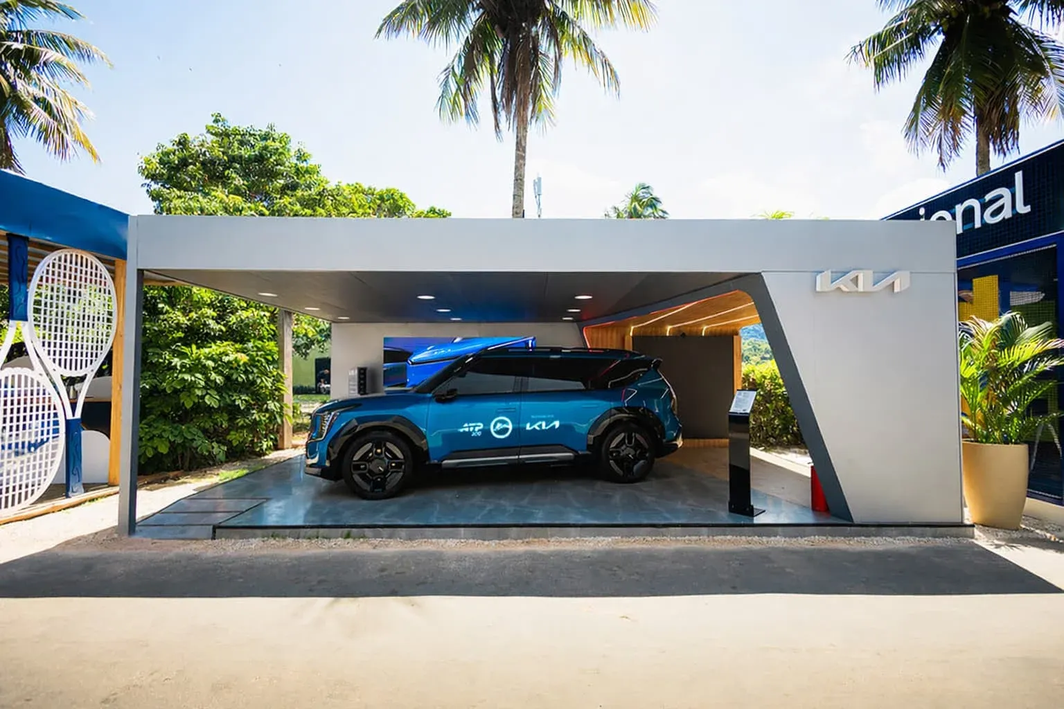 SUV eléctrica Kia exhibida en el pabellón oficial del Rio Open en Brasil, rodeada de palmeras y branding del torneo